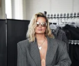 Kobieta w eleganckim szarym garniturze w prążki, stojąca w showroomie modowym. Ma blond włosy, okulary przeciwsłoneczne i srebrny naszyjnik. W tle widoczne są wieszaki z ubraniami i czarne parawany.