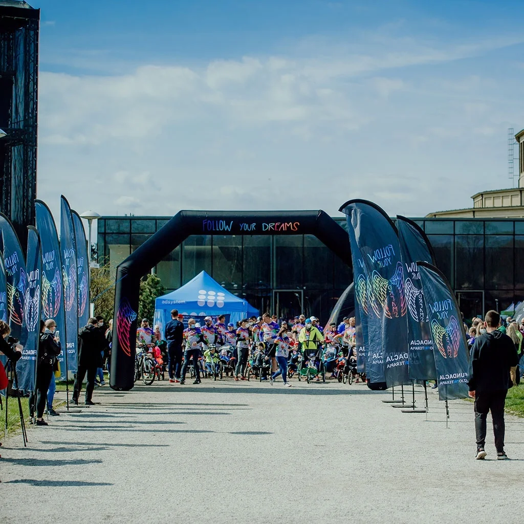 file-meta-description.outdoorsportseventwithbrandedadvertisingflags,aninflatablearch,andabluepromotionaltent,withparticipantsgatheringatthestartingzone.