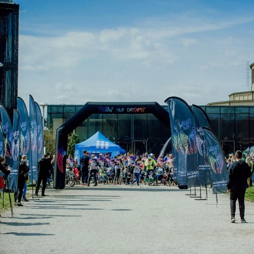 Outdoor sports event with branded advertising flags, an inflatable arch, and a blue promotional tent, with participants gathering at the starting zone.