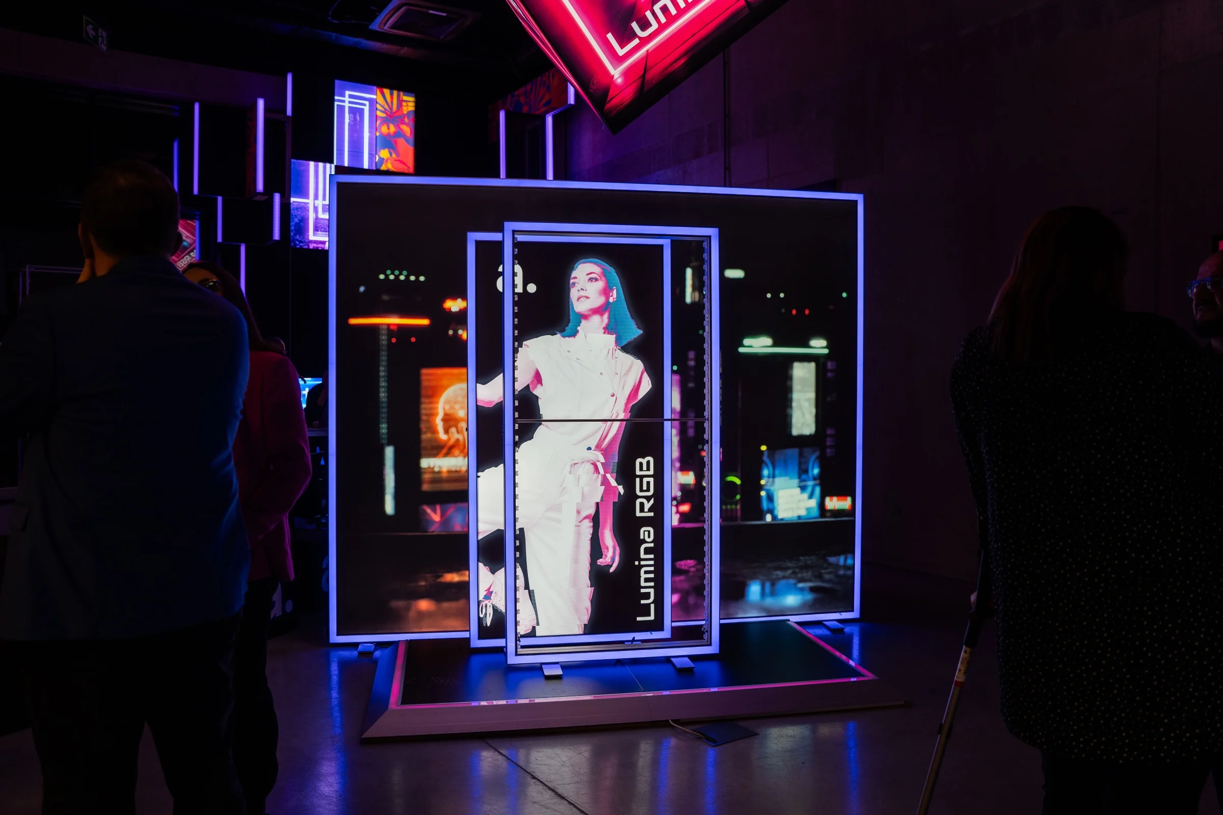 Ekspozycja systemu reklamowego adFrame Lumina RGB w ciemnym, nowoczesnym wnętrzu, z podświetlaną grafiką i elementami neonowymi, przedstawiająca kobietę w jasnym stroju na tle miejskich świateł, otoczona oglądającymi uczestnikami wydarzenia.
