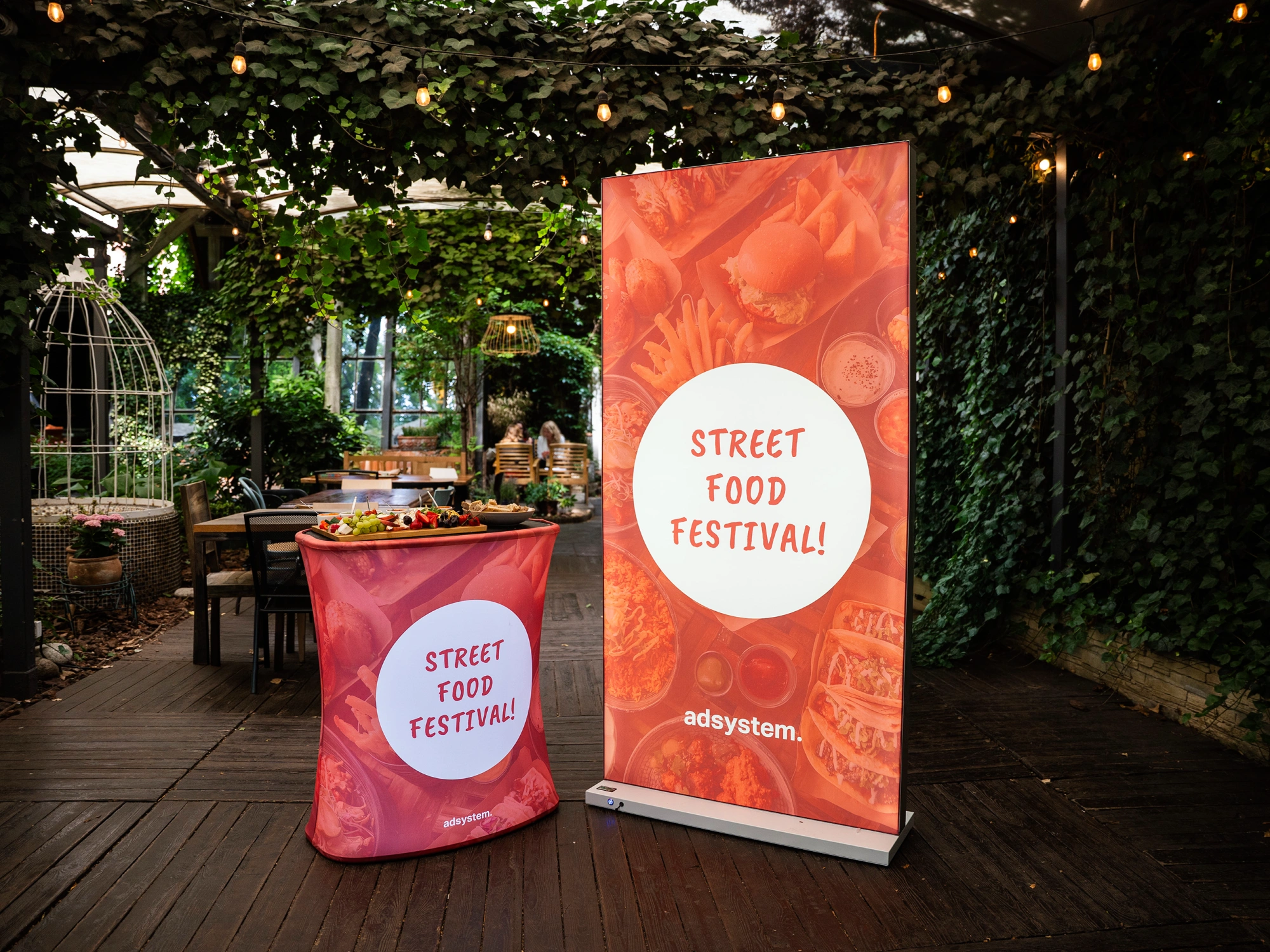 Trybunka i ścianka reklamowa z motywem „Street Food Festival!” ustawione w ogrodowej przestrzeni eventowej pod dekoracyjnymi lampkami.