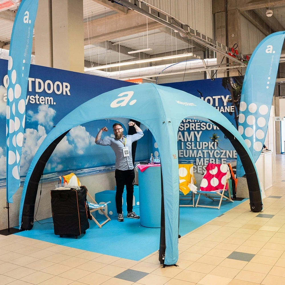Niebieski namiot reklamowy adsystem na hali targowej z mężczyzną i materiałami promocyjnymi w środku.
