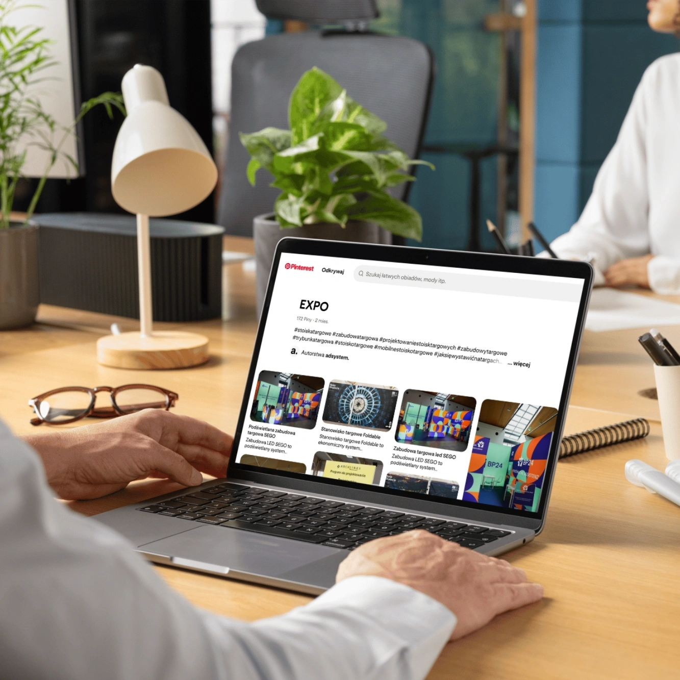 Persona che consulta ispirazioni per stand fieristici sulla piattaforma Pinterest su un laptop, seduta a una scrivania in un ufficio moderno con piante e lampada da tavolo.