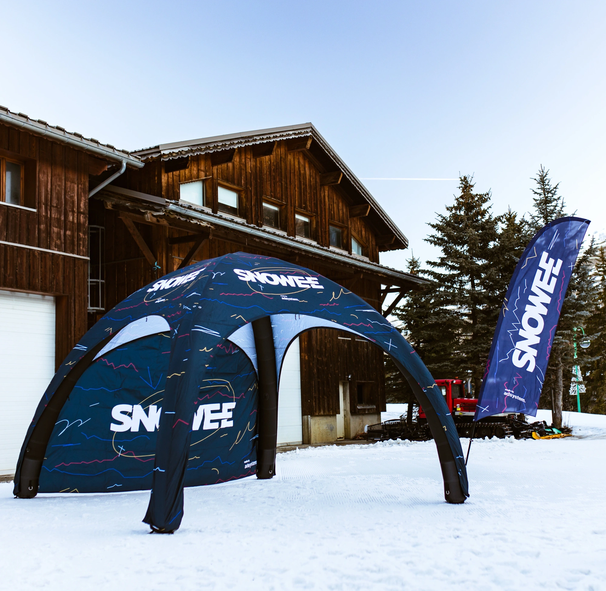 Carpa publicitaria inflable y bandera de Snowee durante el evento Snowee Music Trip 2024 en los Alpes franceses, con productos promocionales de adsystem visibles sobre la nieve.