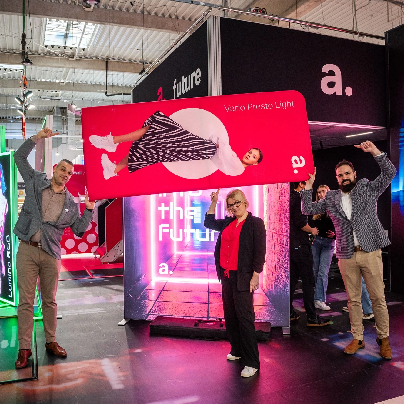 Grupa osób na stoisku targowym adsystem prezentuje lekką ściankę tekstylną Vario Presto Light, trzymając ją uniesioną nad głowami.