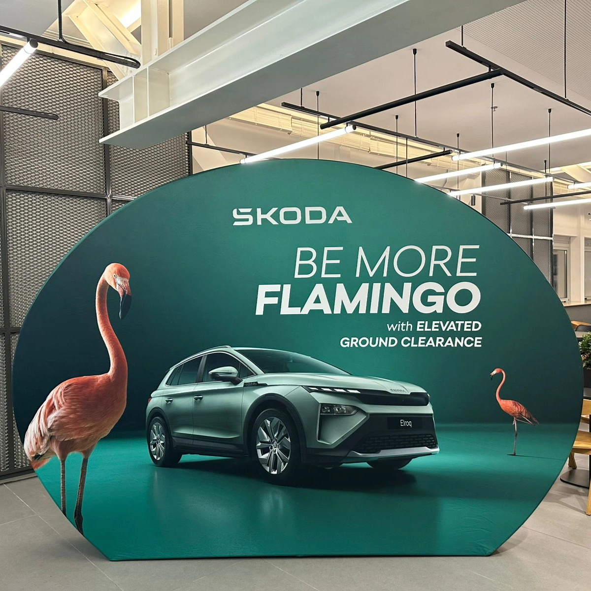 Owalna ścianka tekstylna promująca model Škoda Elroq z hasłem „Be more flamingo”, prezentująca auto na tle flamingów w nowoczesnym wnętrzu ekspozycyjnym.