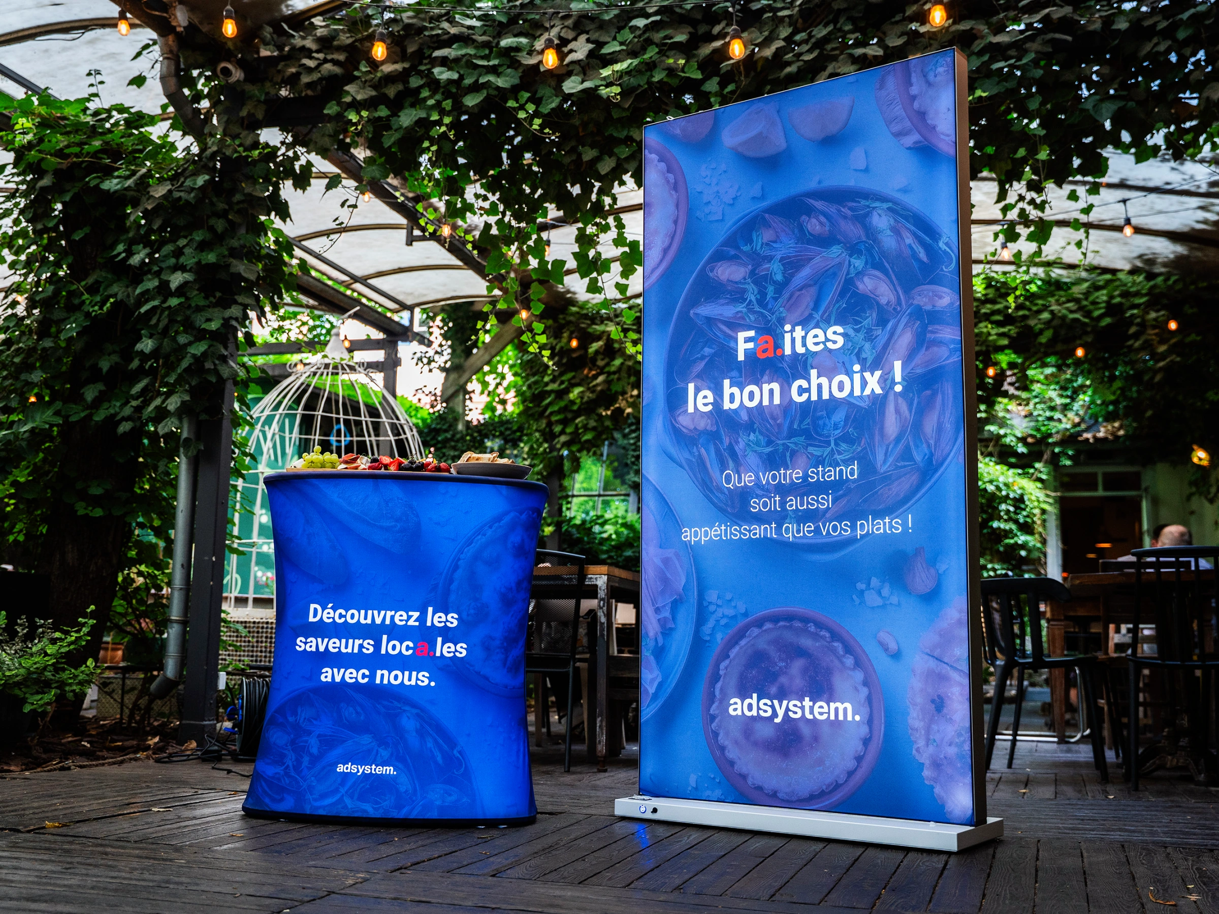 Kaseton podświetlany LED z hasłem „Faites le bon choix !” obok trybunki reklamowej z poczęstunkiem w ogrodzie restauracyjnym.