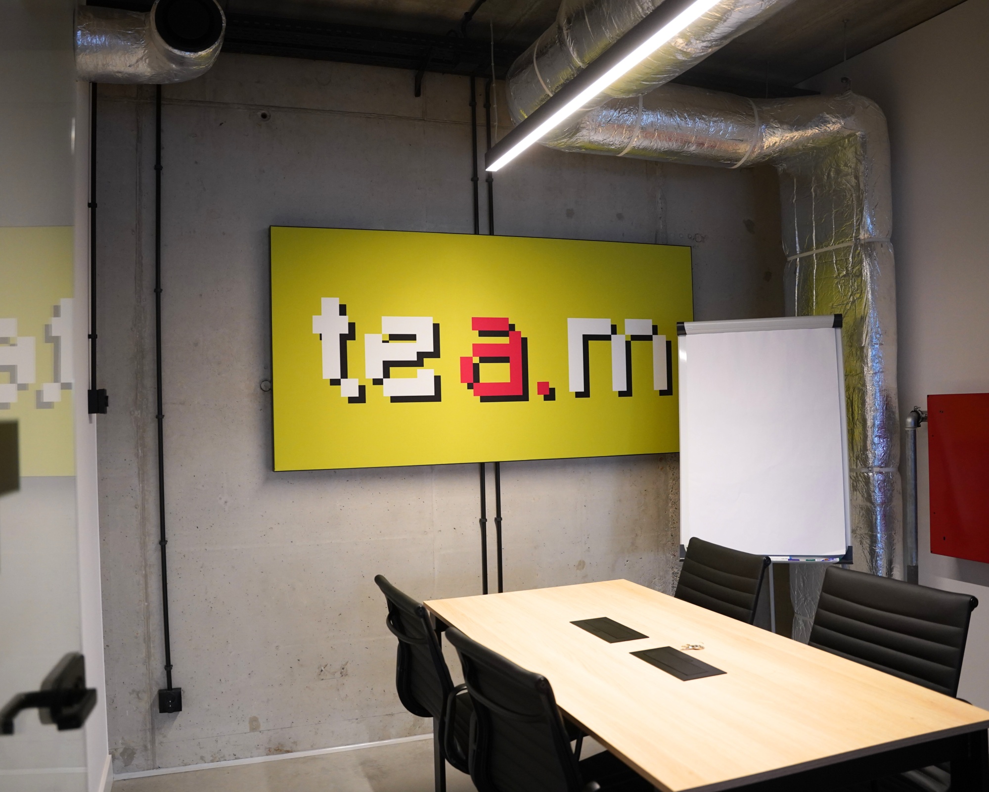 Nowoczesna sala konferencyjna z żółtym kasetonem LED na ścianie, przedstawiającym napis w stylu pikselowym, obok tablica flipchart i stół z czarnymi krzesłami. Zapytaj ChatGPT