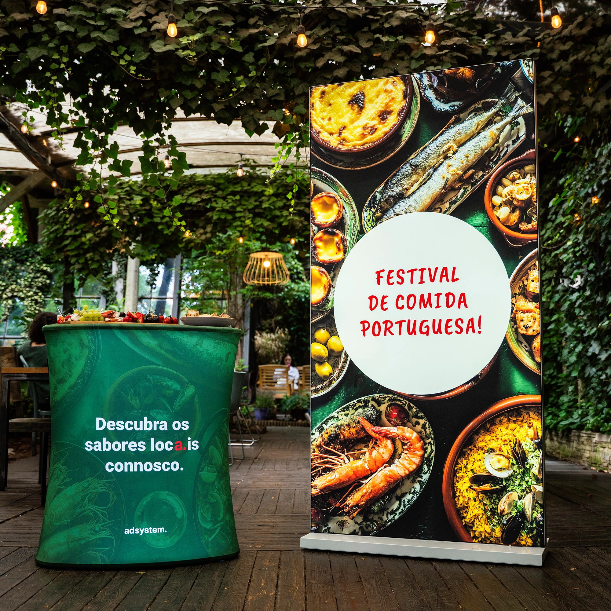 Trybunka i ścianka reklamowa adsystem w zielonej kolorystyce z portugalskimi hasłami „Descubra os sabores locais connosco” i „Festival de comida portuguesa!”, ustawione w ogrodowej przestrzeni z dekoracyjnymi lampkami.