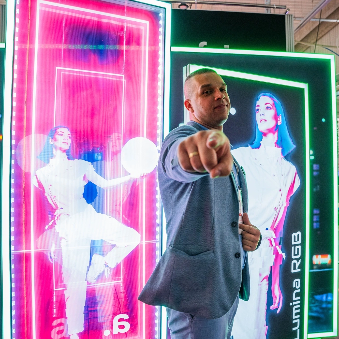 Hombre con chaqueta gris señalando a la cámara frente a paneles publicitarios LED Lumina RGB iluminados.