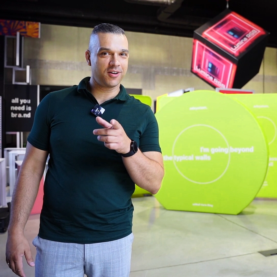 Mężczyzna w zielonej koszulce wskazuje w stronę kamery, stojąc na tle kolorowych systemów wystawienniczych.