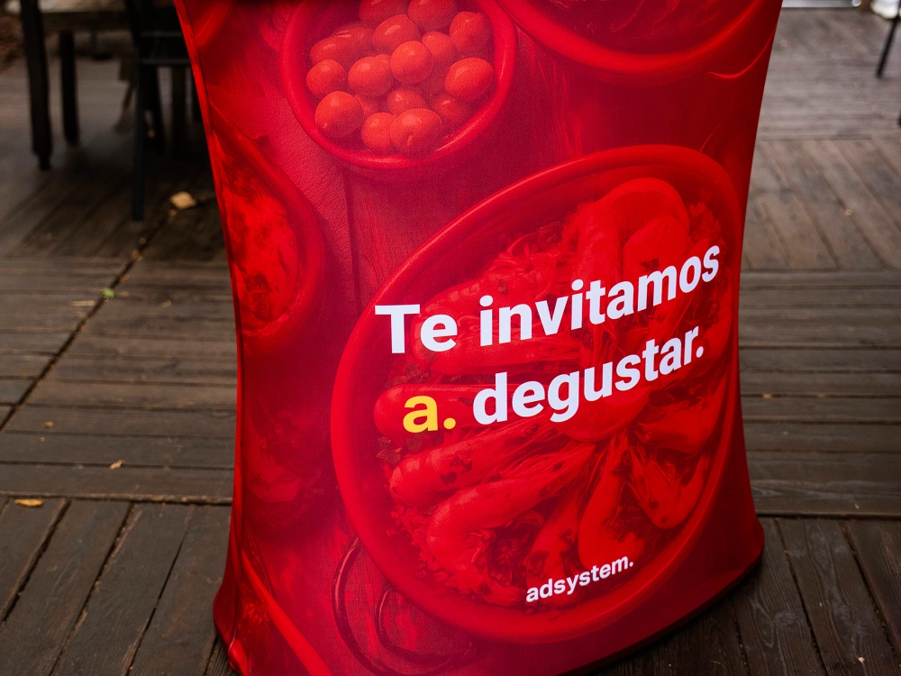 Zbliżenie na trybunkę reklamową adsystem w czerwonej kolorystyce z hiszpańskim hasłem „Te invitamos a degustar.” nadrukowanym na froncie.