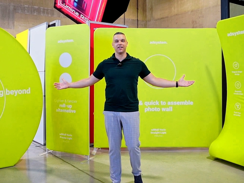 Una persona posa con los brazos abiertos frente a un photocall textil Vario Straight Light verde de adsystem instalado en un showroom.