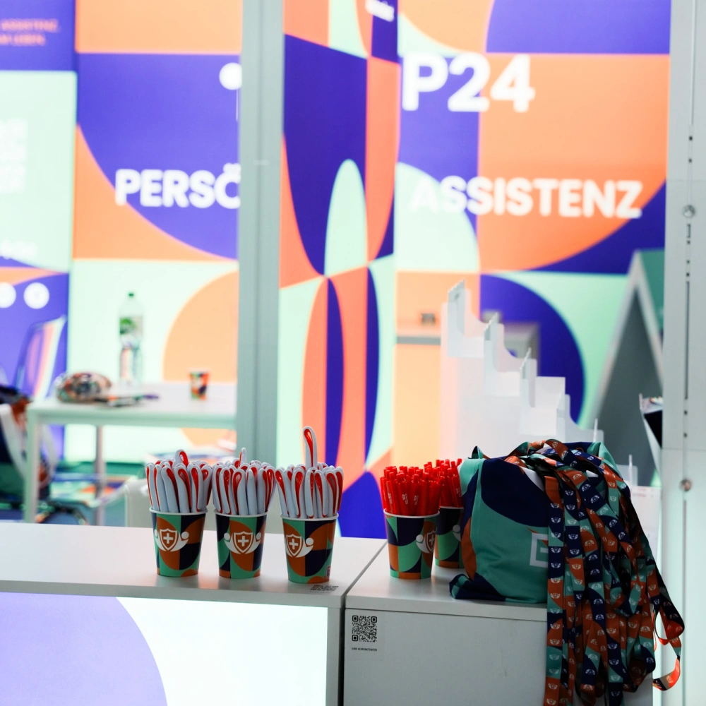 Stoisko targowe BP24 z kolorową podświetlaną zabudową w tle, wyposażone w gadżety promocyjne takie jak długopisy, smycze i kubeczki z nadrukiem firmowym.