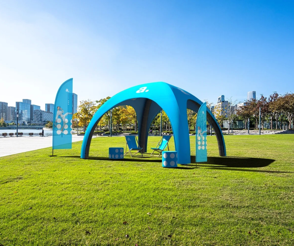 Niebieski namiot reklamowy pneumatyczny z leżakami i flagami plażowymi ustawiony na trawniku w parku miejskim z widokiem na nowoczesne wieżowce.