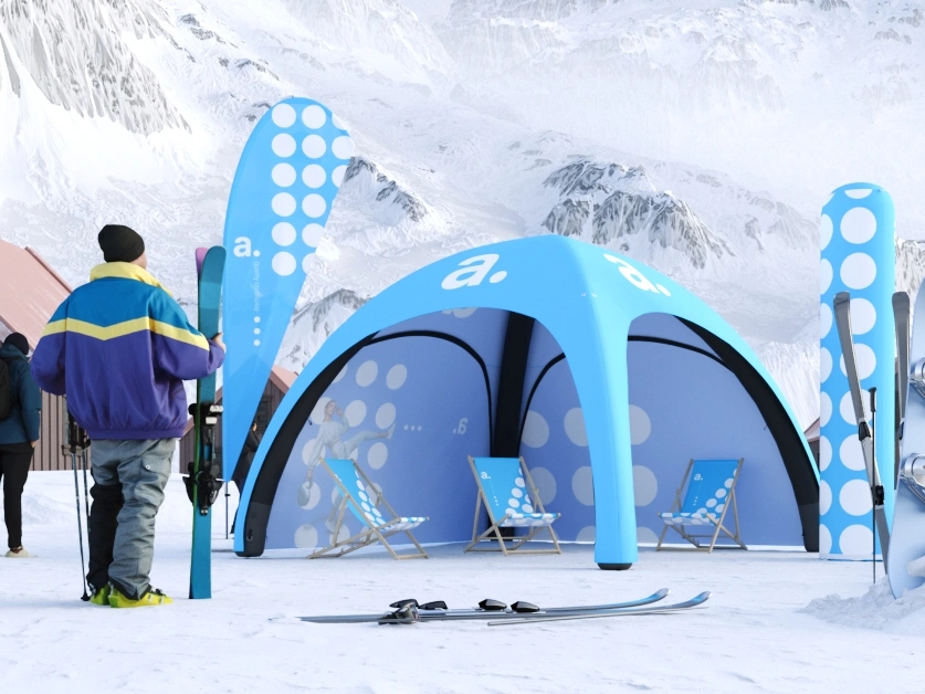 Zimowa strefa promocyjna adsystem z niebieskim namiotem pneumatycznym, leżakami i flagami reklamowymi na tle ośnieżonych gór.