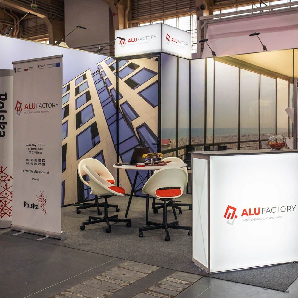 Stoisko targowe Alu Factory z podświetlaną ladą, roll-upem informacyjnym, grafikami przedstawiającymi nowoczesne fasady budynków oraz strefą rozmów wyposażoną w stolik i krzesła.