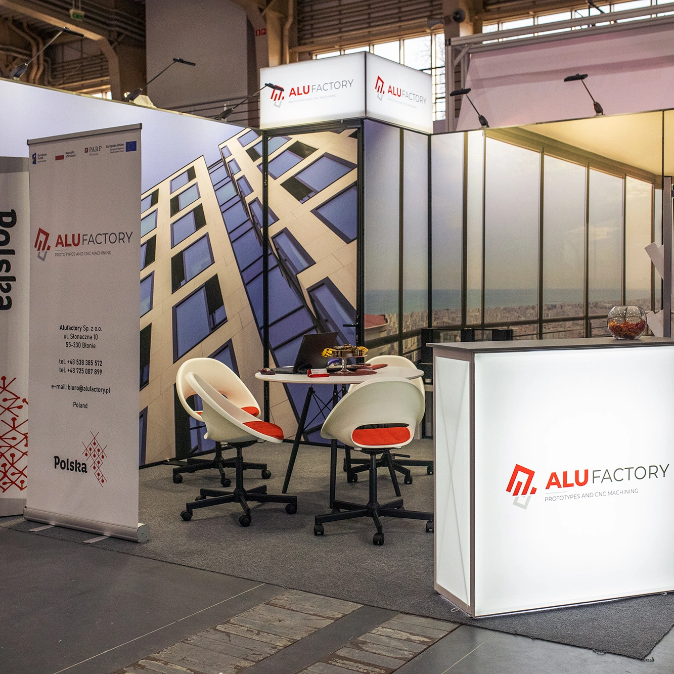 Stoisko targowe Alu Factory z podświetlaną ladą, roll-upem informacyjnym, grafikami przedstawiającymi nowoczesne fasady budynków oraz strefą rozmów wyposażoną w stolik i krzesła.