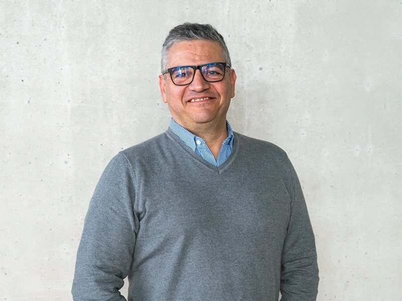 Mężczyzna w okularach i szarym swetrze pozuje na tle jasnej betonowej ściany, uśmiechając się do obiektywu.