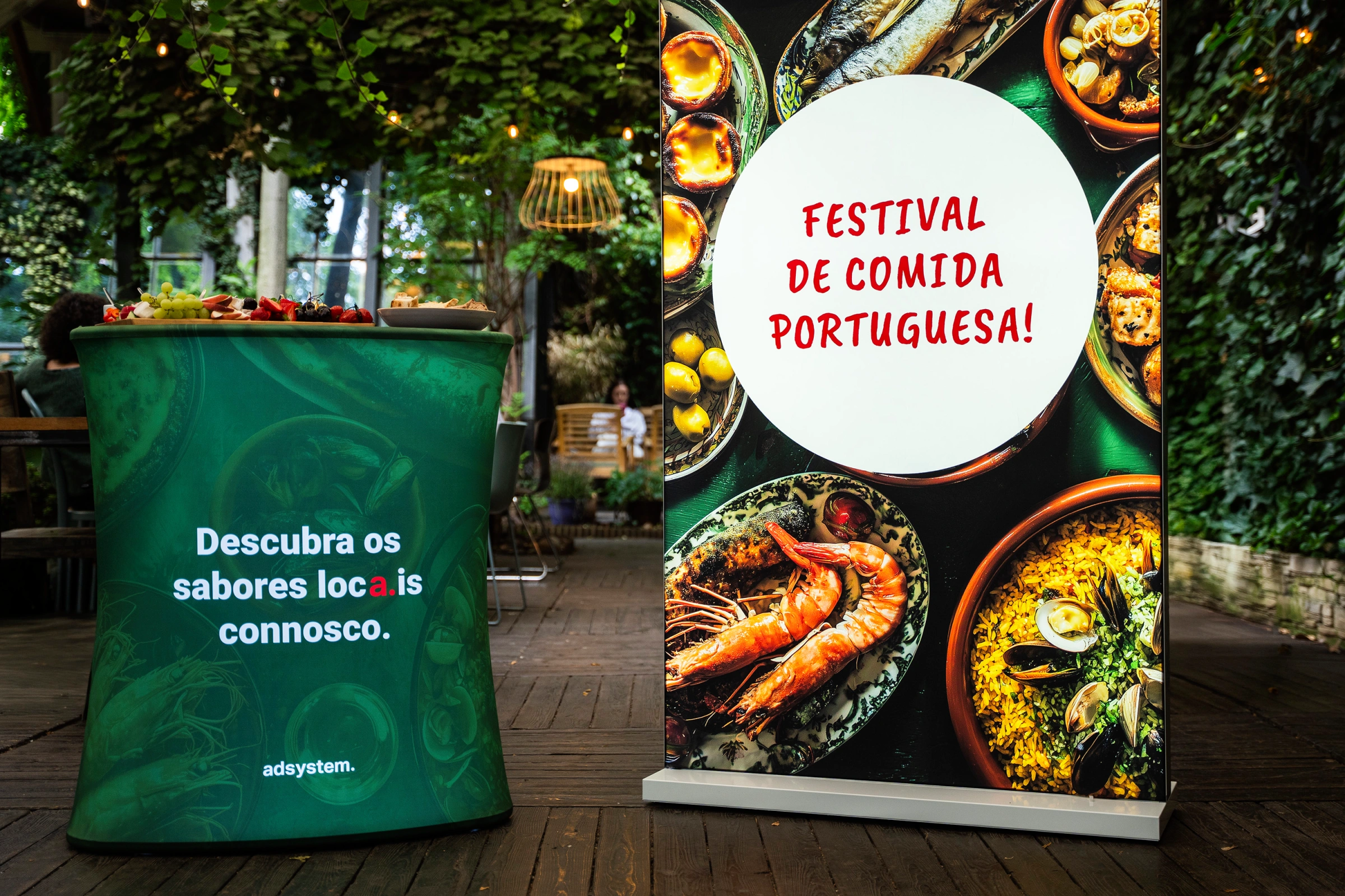 Trybunka i ścianka reklamowa adsystem w zielonej oprawie z portugalskimi hasłami „Descubra os sabores locais connosco” i „Festival de comida portuguesa!”, ustawione w ogrodowej przestrzeni eventowej.
