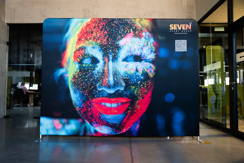Photocall textil publicitario para ferias con estructura modular de aluminio y gráfica impresa de alta resolución que muestra un rostro iluminado con pintura fluorescente.