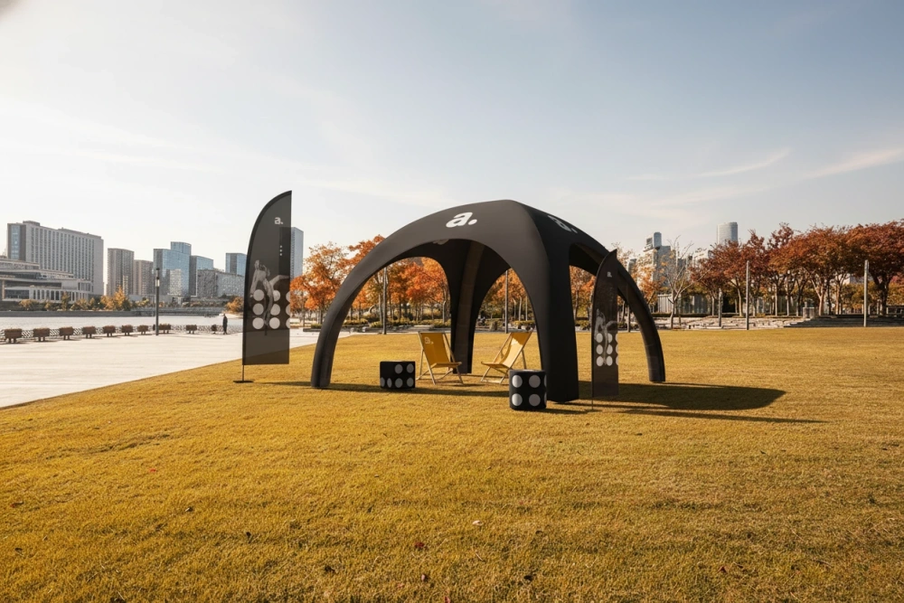 Czarny nadmuchiwany namiot eventowy z flagami reklamowymi i leżakami, ustawiony na jesiennym trawniku z panoramą miasta.