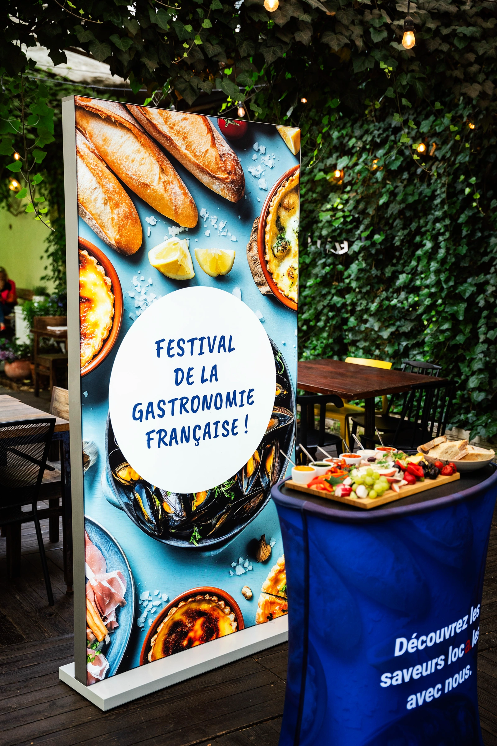 Kaseton reklamowy LED z grafiką „Festival de la Gastronomie Française” obok trybunki z przekąskami w ogrodzie restauracyjnym.