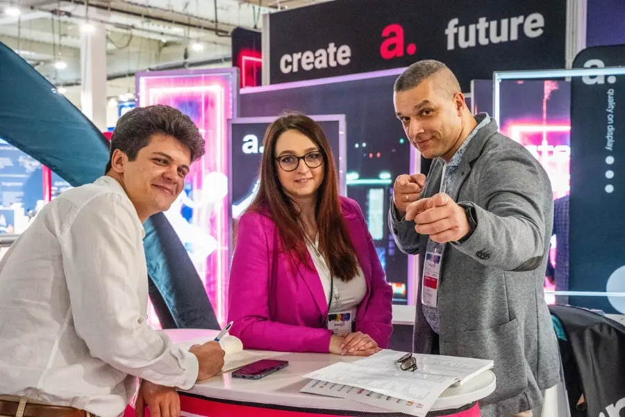 Trzy osoby przy stoisku adsystem – uśmiechnięci uczestnicy targów rozmawiają i pozują do zdjęcia.