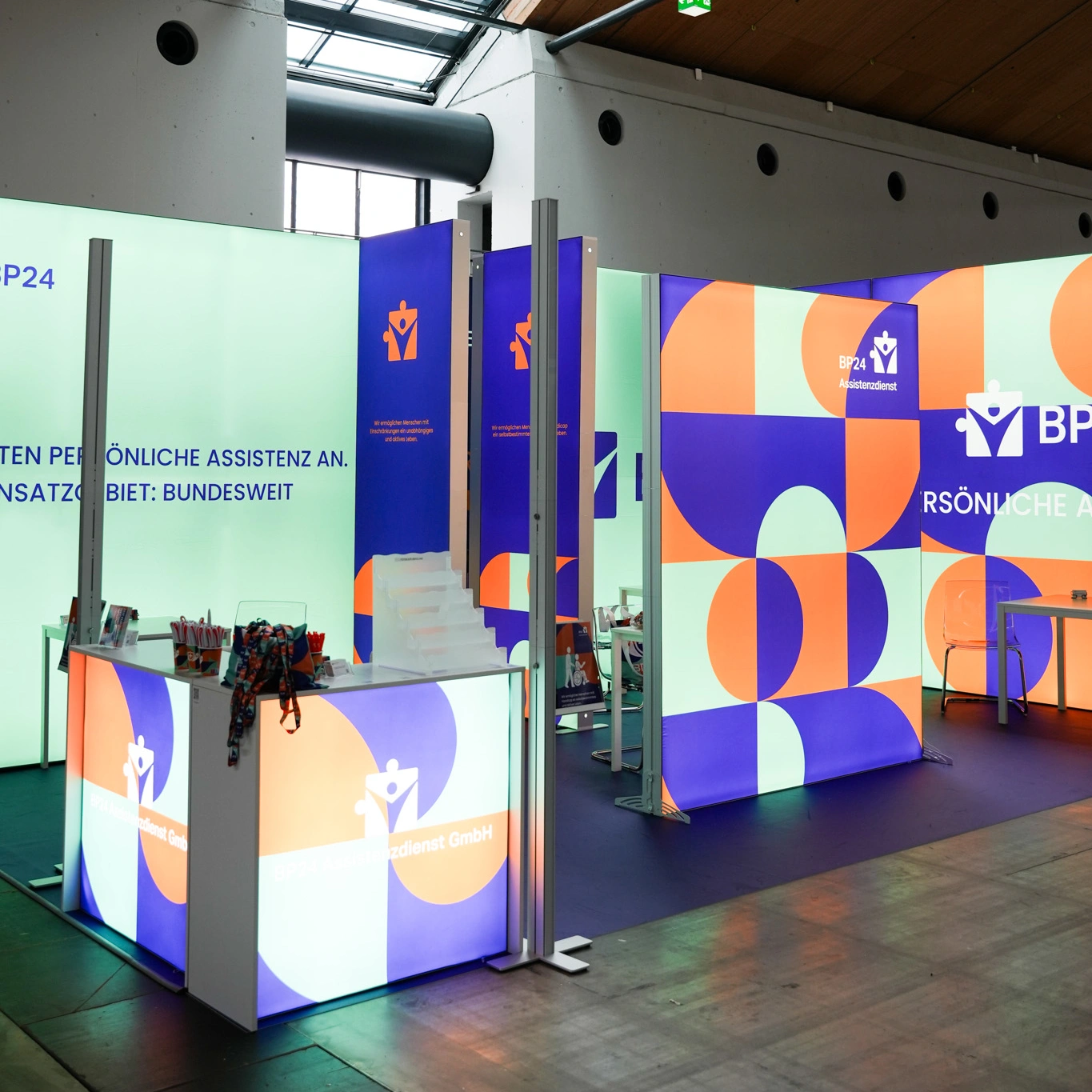 Stoisko targowe BP24 z podświetlaną zabudową w geometryczne wzory w kolorach pomarańczowym, fioletowym i miętowym, wyposażone w ladę, stojak na ulotki oraz strefę rozmów.