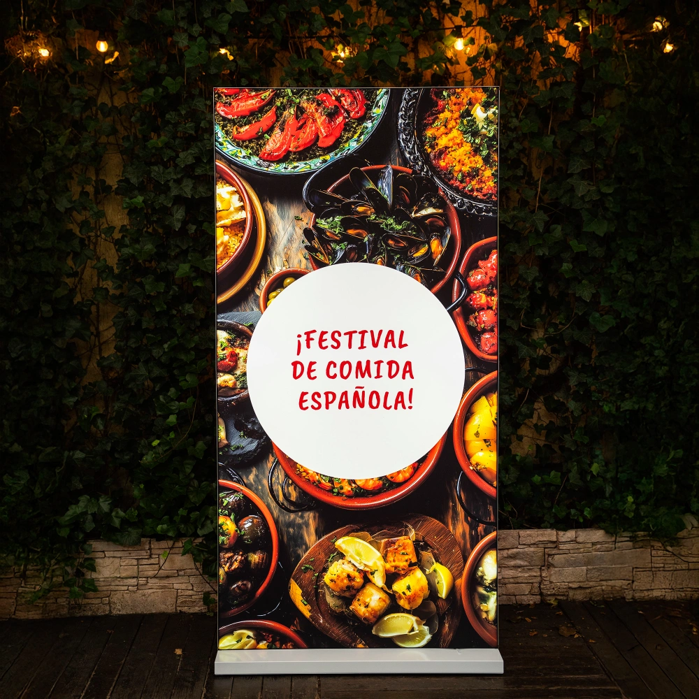 Ścianka reklamowa adsystem z nadrukiem „¡Festival de comida española!” przedstawiająca hiszpańskie tapas, ustawiona na tle zielonej ściany z bluszczu i lampek dekoracyjnych.
