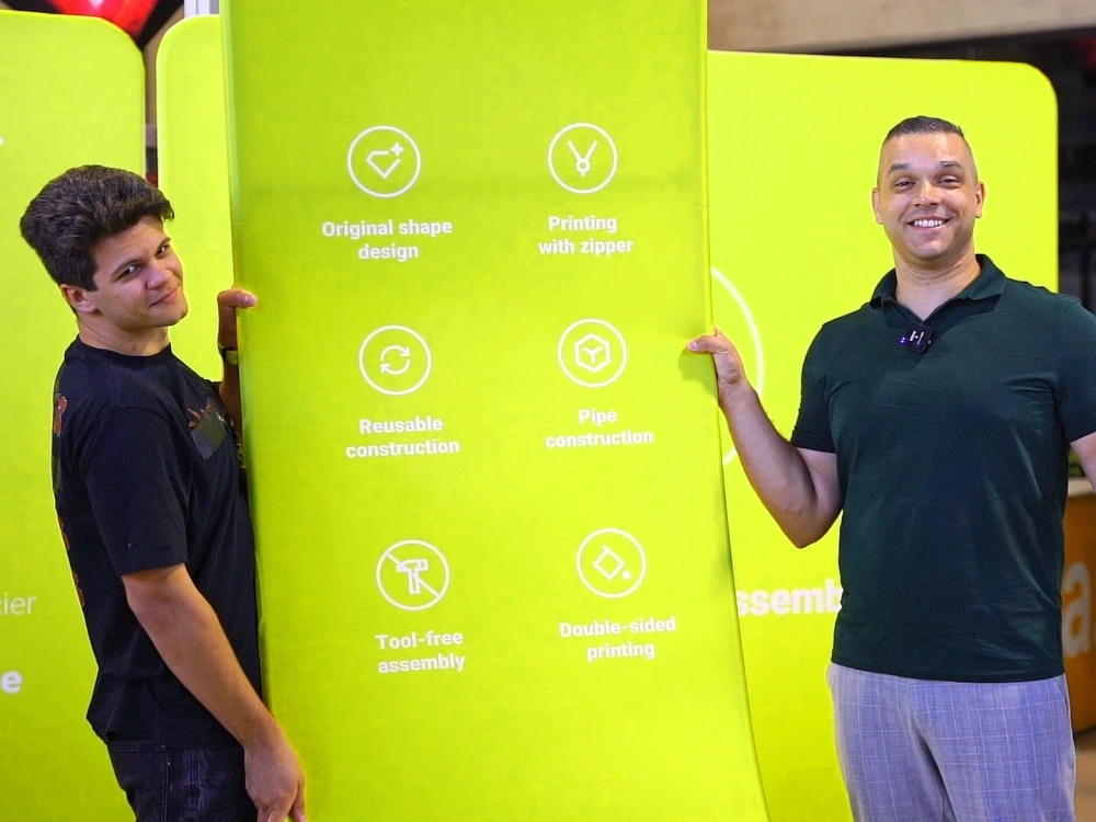 Dos personas sostienen un photocall con forma curva Vario S de color verde en un showroom, mostrando los iconos que explican sus características principales como montaje sin herramientas y estructura reutilizable.