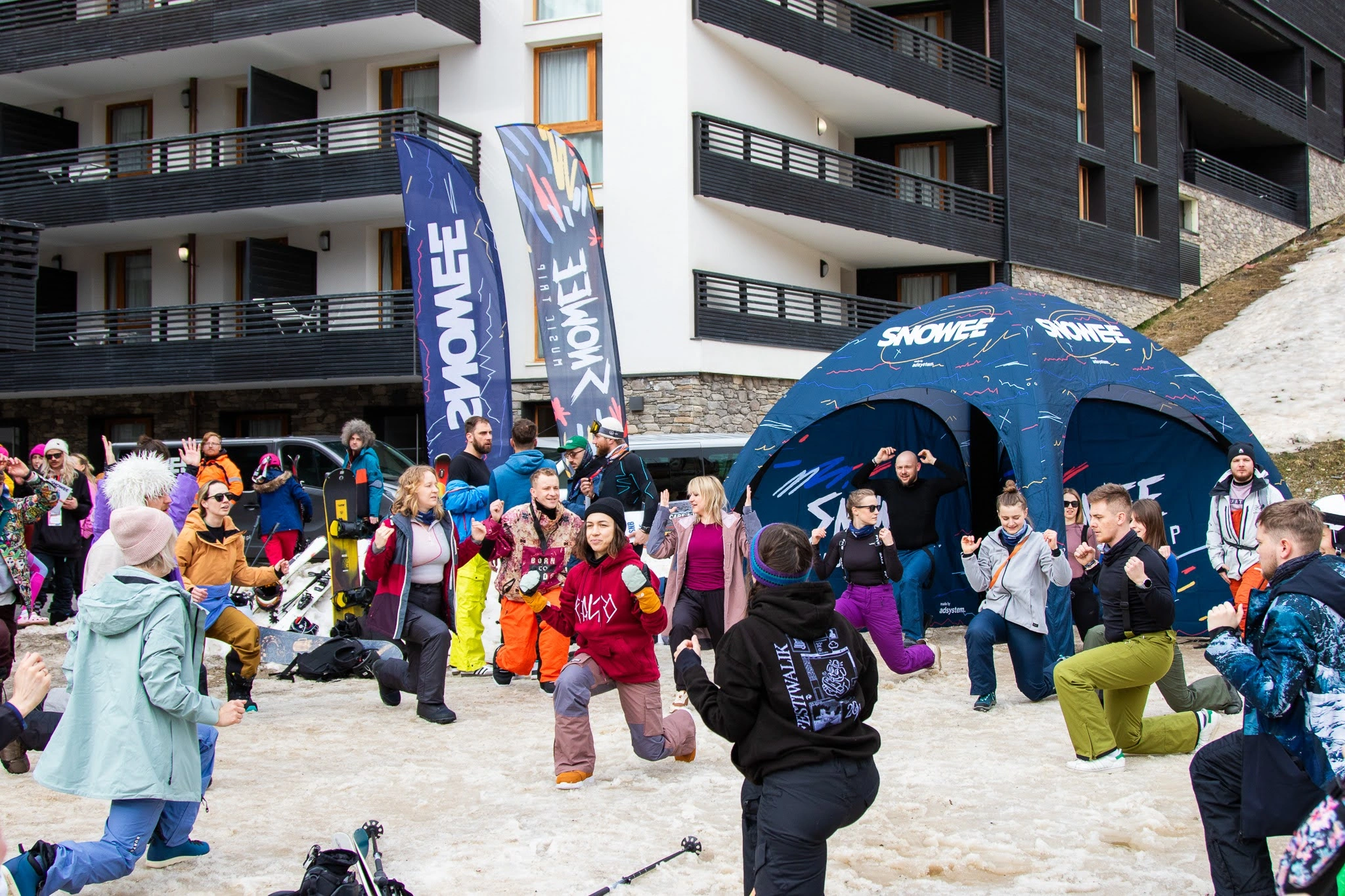 Grupa uczestników zimowego eventu przed hotelem ćwiczy przy namiocie i flagach reklamowych SNOWEE, oznaczonych logo adsystem.