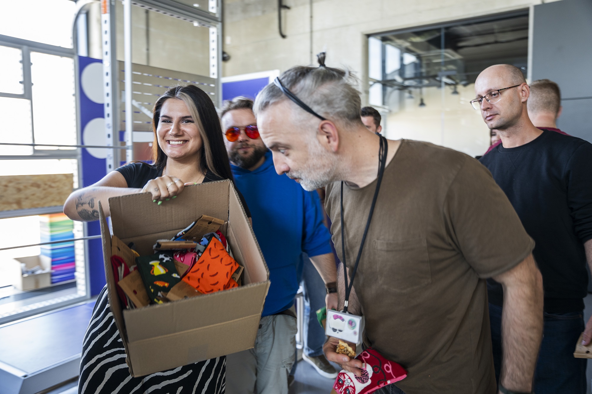 Uśmiechnięta kobieta trzyma karton z kolorowymi skarpetkami, otoczona przez współpracowników w biurze – moment integracji i wspólnej akcji pracowniczej.