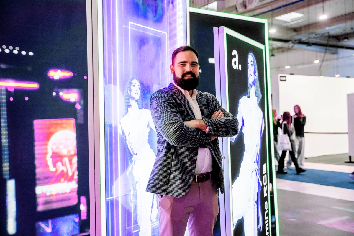 Mężczyzna pozuje na tle podświetlanych paneli RGB z grafiką promocyjną, prezentując nowoczesne rozwiązania wizualne na stoisku targowym.