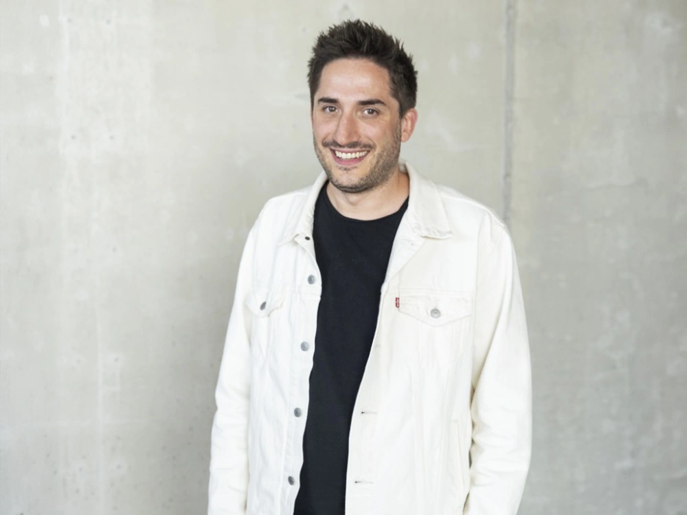 Homme souriant portant une veste claire et un t-shirt noir, debout devant un mur en béton.