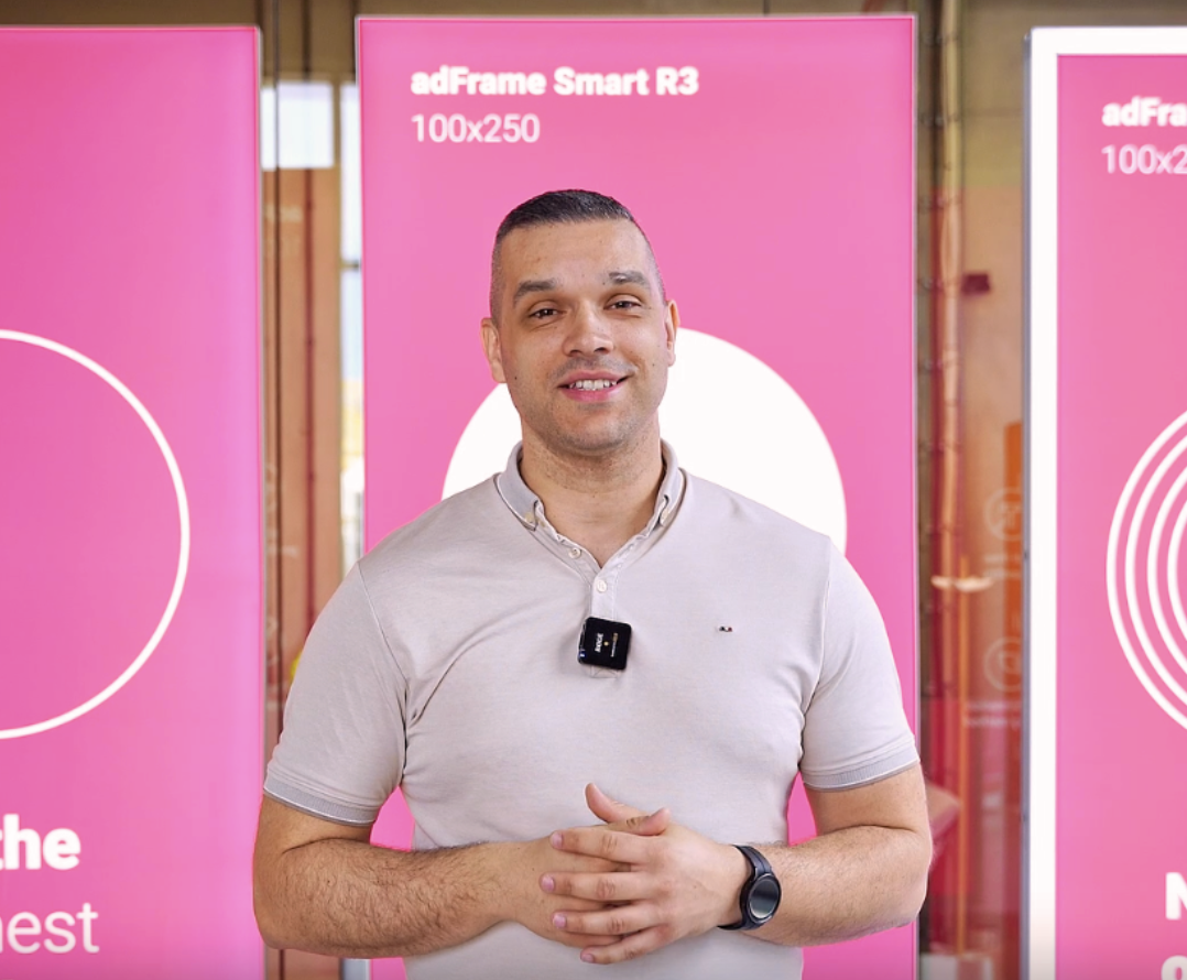 Mężczyzna prezentujący kasetony adFrame Smart R3 100x250 na tle różowych paneli podczas nagrania w showroomie lub na targach.
