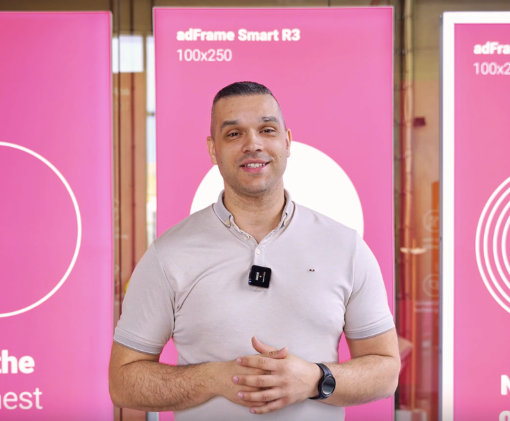 Mężczyzna prezentujący kasetony adFrame Smart R3 100x250 na tle różowych paneli podczas nagrania w showroomie lub na targach.