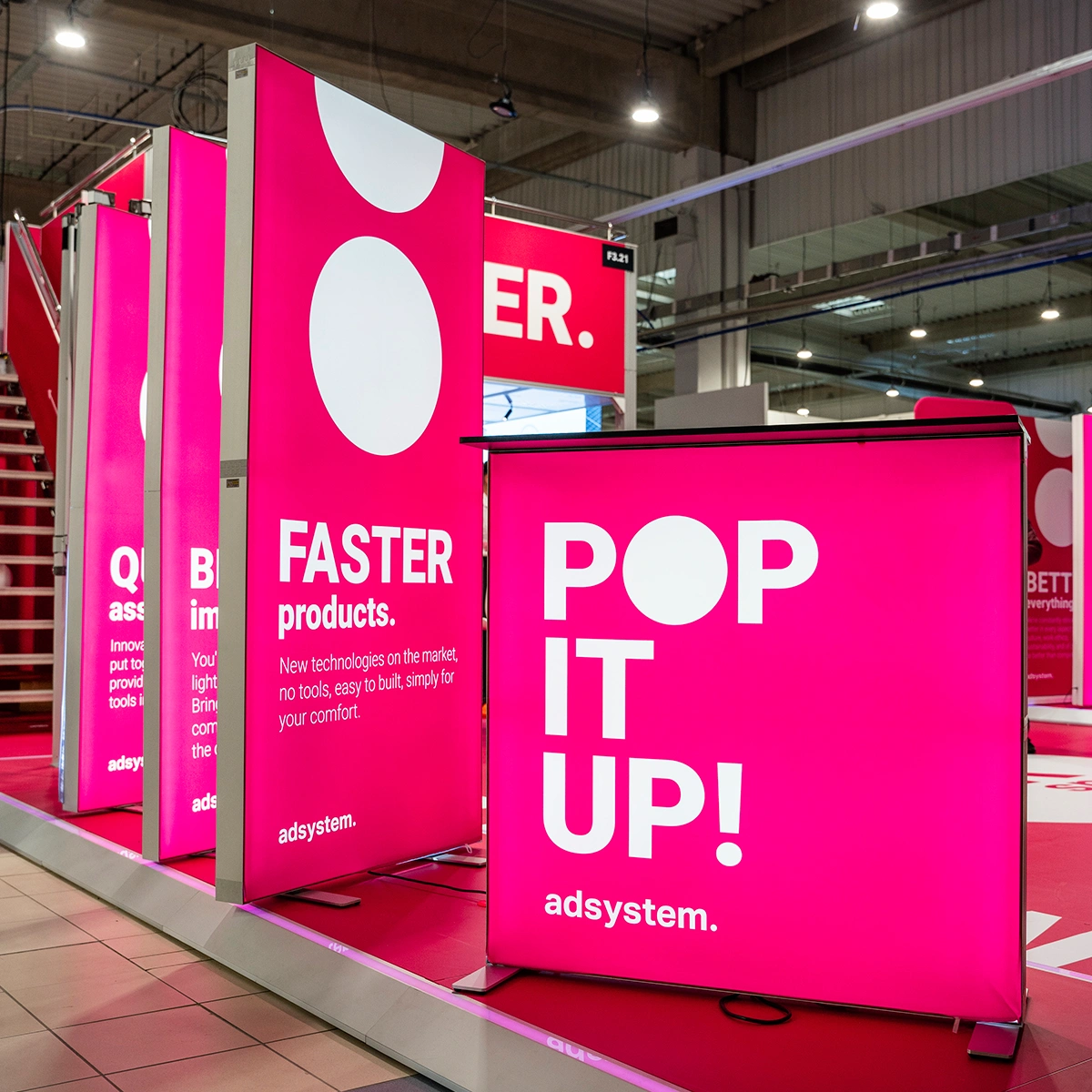 Zestaw podświetlanych kasetonów tekstylnych i trybunek z hasłami „POP IT UP!” i „FASTER products”, intensywny różowy kolor, branding adsystem, ekspozycja targowa w stylu modularnym.