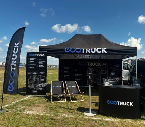 Stoisko sponsorskie EcoTruck na wydarzeniu plenerowym – namiot z nadrukiem, flagi reklamowe, trybunka, leżaki i roll-up z grafiką techniczną ciężarówki, ustawione na otwartym terenie pod niebieskim niebem. 