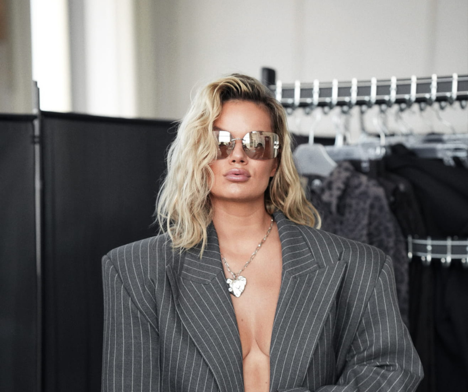 Kobieta w eleganckim szarym garniturze w prążki, stojąca w showroomie modowym. Ma blond włosy, okulary przeciwsłoneczne i srebrny naszyjnik. W tle widoczne są wieszaki z ubraniami i czarne parawany.