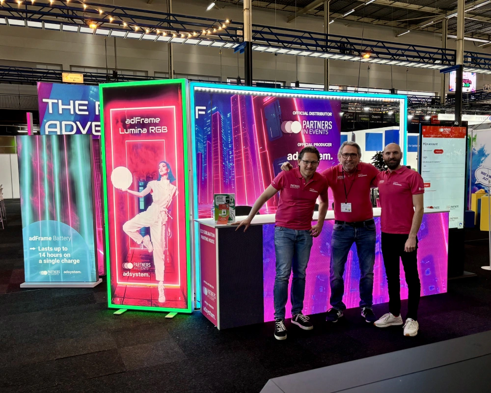 Stoisko adsystem z podświetlanymi kasetonami adFrame i zespołem w różowych koszulkach podczas targów branżowych.