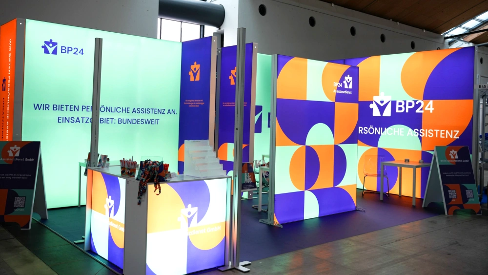 Stoisko targowe BP24 z podświetlaną zabudową w geometryczne wzory w kolorach pomarańczowym, fioletowym i miętowym, wyposażone w ladę z materiałami promocyjnymi, stojaki informacyjne oraz strefę do rozmów.