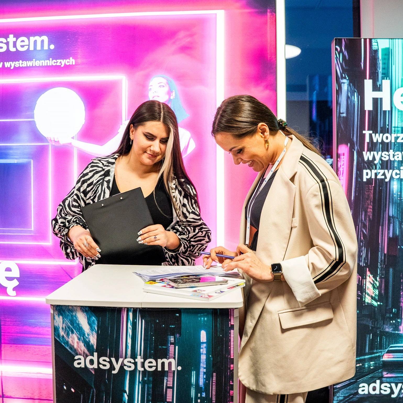 file-meta-description.dwiekobietyrozmawiająprzytrybuncereklamowejadsystem,natlepodświetlanychsystemówwystawienniczychzneonowymigrafikamiihasłami.