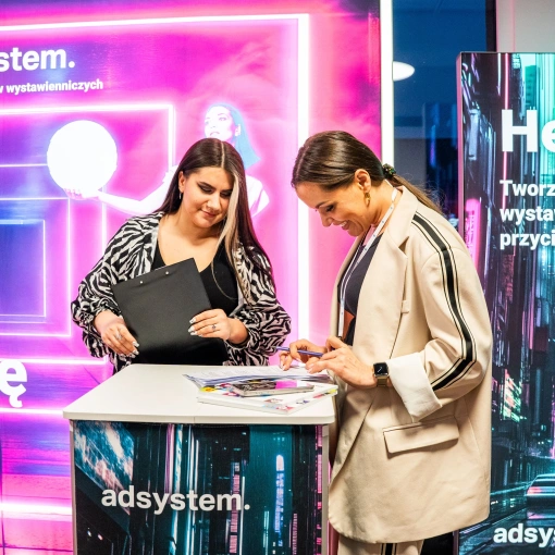 Dwie kobiety rozmawiają przy trybunce reklamowej adsystem, na tle podświetlanych systemów wystawienniczych z neonowymi grafikami i hasłami.