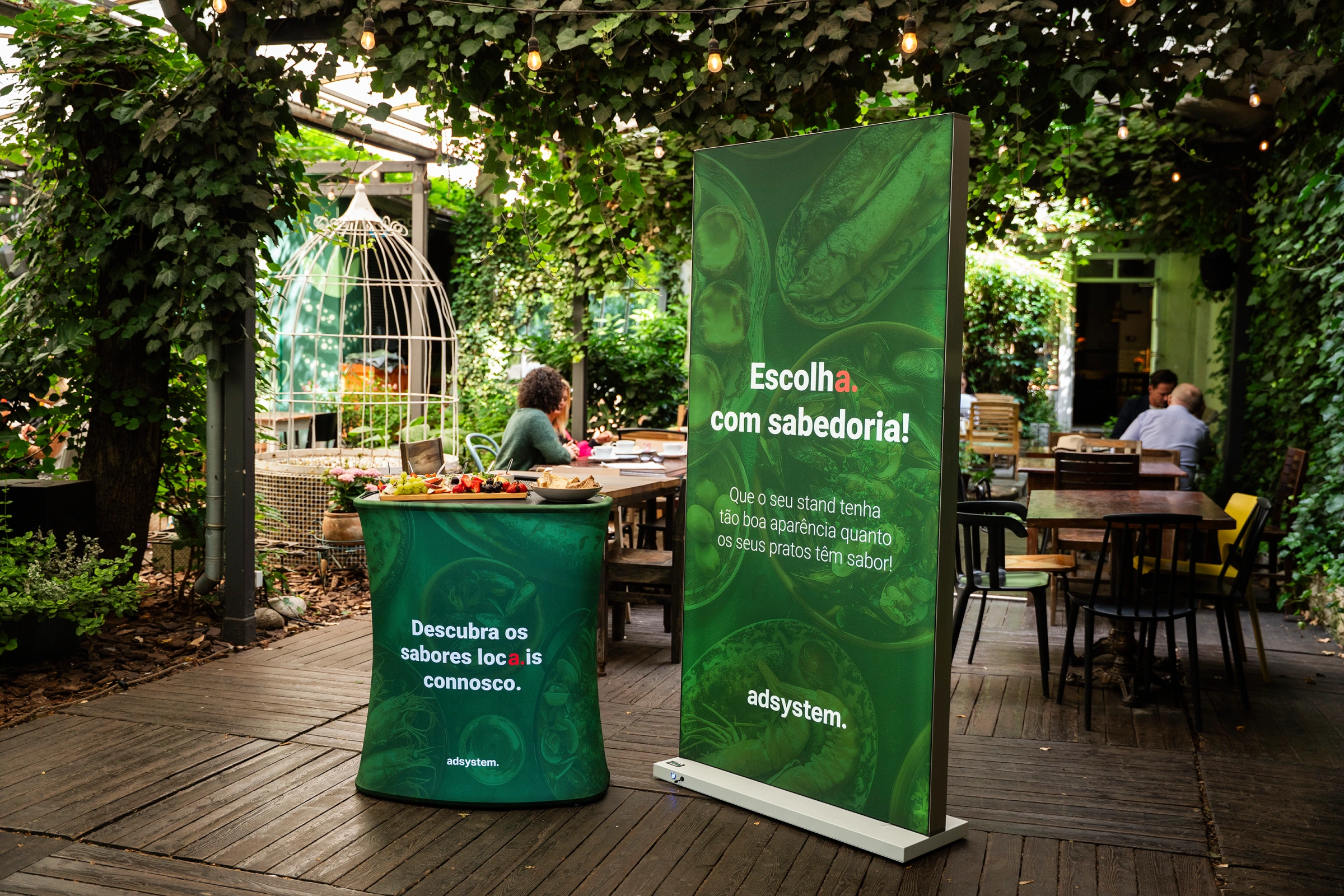 Trybunka i ścianka reklamowa adsystem w zielonej oprawie z portugalskimi hasłami promocyjnymi, ustawione w ogrodowej przestrzeni z dekoracyjnymi lampkami.