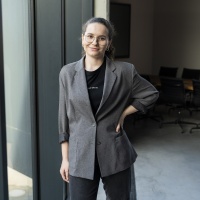 Femme souriante portant des lunettes et une veste grise, debout près d’une baie vitrée dans un bureau moderne.