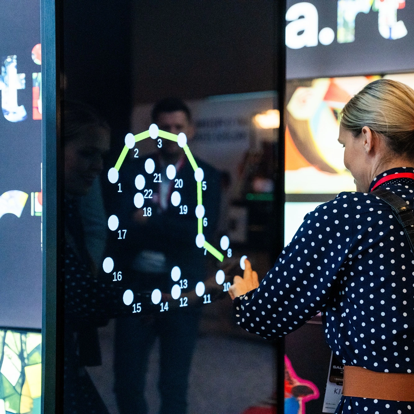 Osoba korzystająca z infokiosku na targach, rysując linię po ponumerowanych punktach na dużym ekranie.