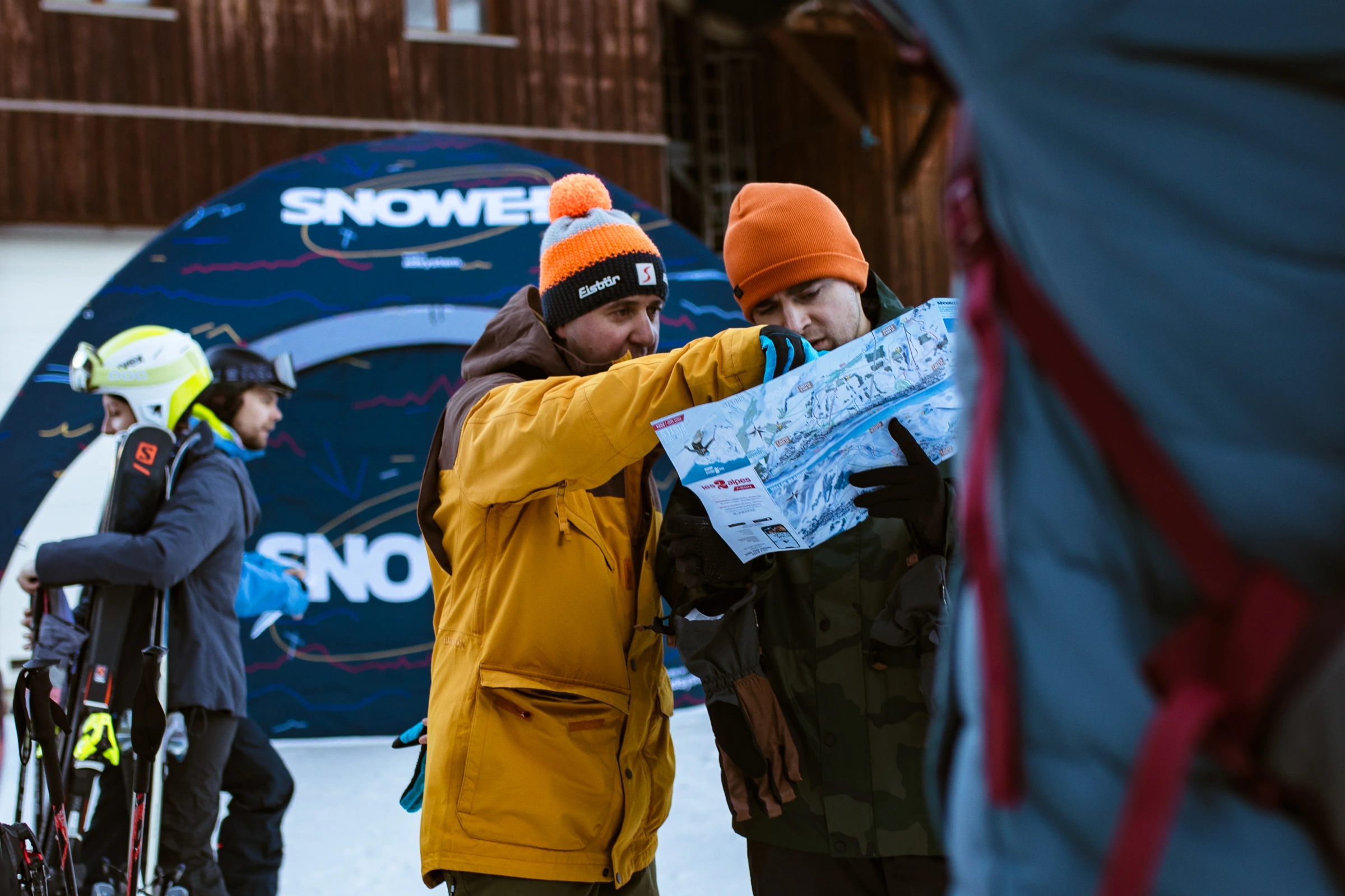 Dwie osoby w zimowych kurtkach przeglądają mapę ośrodka narciarskiego, na tle namiotu reklamowego z logo Snowee.