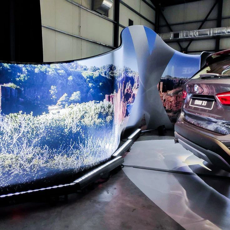 Podświetlana ścianka tekstylna o falistej formie, przedstawiająca krajobraz natury, ustawiona obok samochodu BMW X1 w przestrzeni wystawienniczej, tworząc efektowną scenografię promocyjną.
