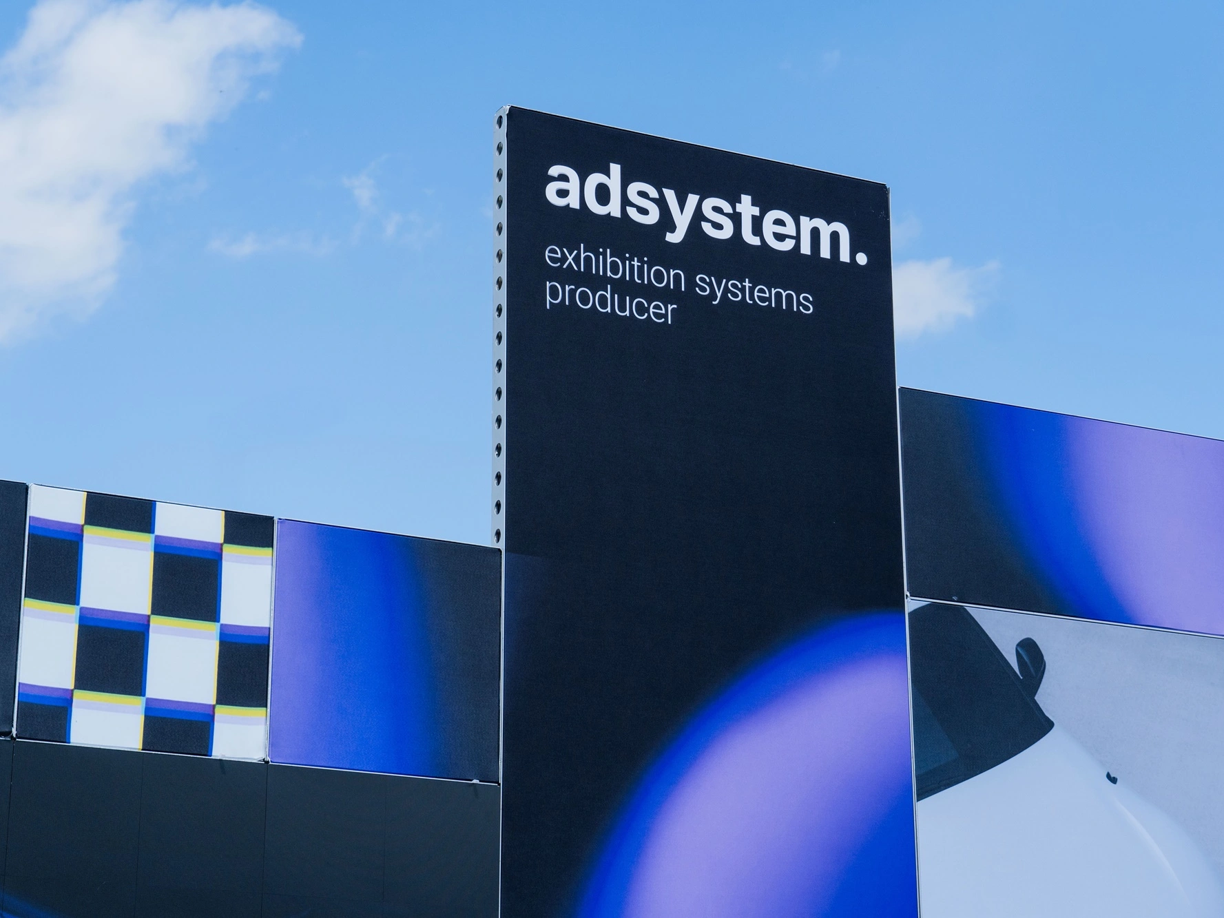 Fragment stoiska adsystem z widocznym logo i hasłem „exhibition systems producer” na tle nieba podczas wydarzenia Ultrace 2025.
